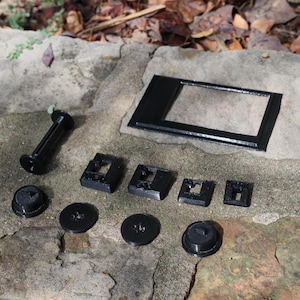 May include: An assortment of black 3D-printed components, including a rectangular frame, several square and circular pieces, and a cylindrical handle. The items are arranged on a rough, stone surface.