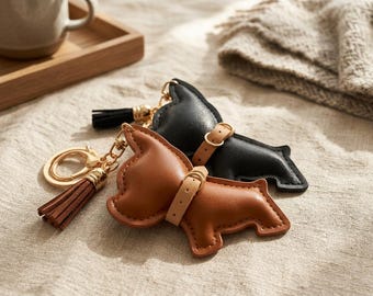 May include: Two dog-shaped keychains made of smooth leather, one in black and one in tan. Each keychain features a gold-tone metal ring, a matching leather tassel, and a small, light-colored leather collar detail around the neck area.