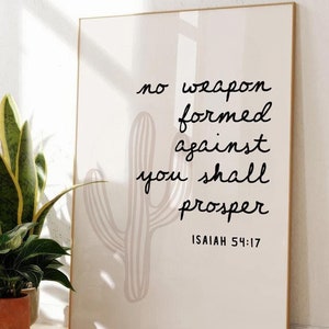 Puede incluir: Una impresión enmarcada blanca con una ilustración de cactus y el texto "no weapon formed against you shall prosper Isaiah 54:17". La impresión está colgada en una pared blanca en una habitación con plantas.
