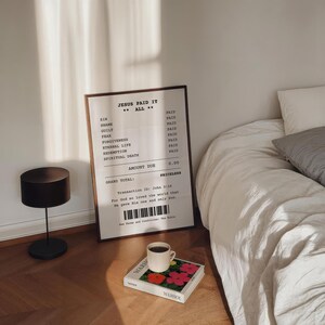 May include: A framed print with a receipt-style design that reads "Jesus Paid It All." The print is on a white background with black text. The print is hanging on a white wall above a bed with white linens. A black lamp is on the floor next to the print. A book with a colorful cover is on a table in front of the bed.