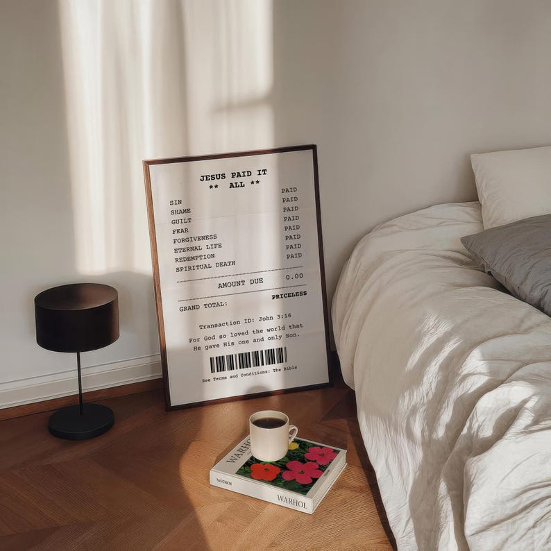May include: A framed print with a receipt-style design that reads "Jesus Paid It All." The print is on a white background with black text. The print is hanging on a white wall above a bed with white linens. A black lamp is on the floor next to the print. A book with a colorful cover is on a table in front of the bed.