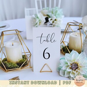 May include: A white card with black text reading "Table 6" sits on a gold geometric stand. The stand holds a white candle. The card is on a table with a white tablecloth. There are flowers and other candles in the background.