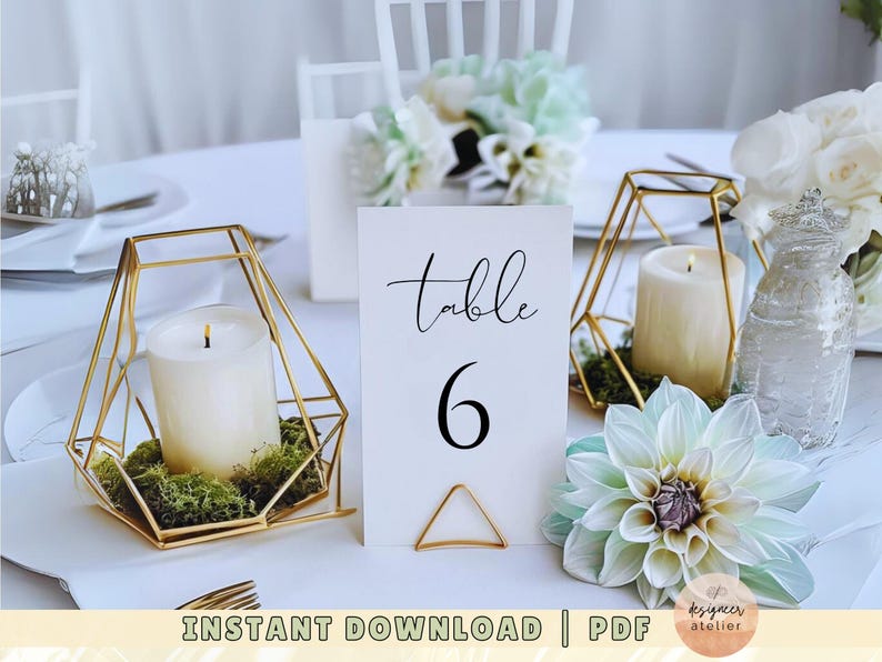 May include: A white card with black text reading "Table 6" sits on a gold geometric stand. The stand holds a white candle. The card is on a table with a white tablecloth. There are flowers and other candles in the background.