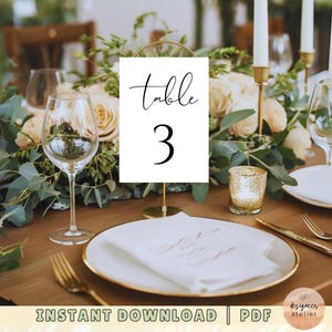May include: White table number card with black calligraphy that reads "Table 3" on a gold stand. The card is on a wooden table with a white napkin, gold silverware, and a gold candle holder.