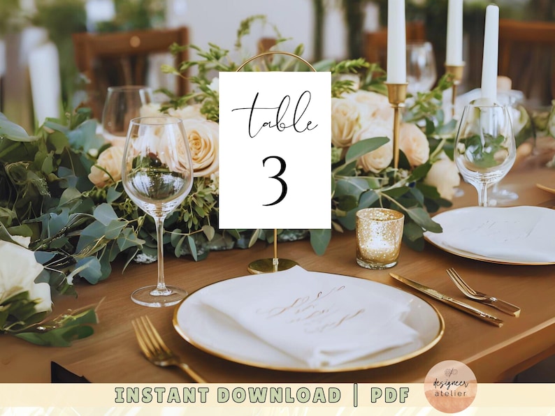 May include: White table number card with black calligraphy that reads "Table 3" on a gold stand. The card is on a wooden table with a white napkin, gold silverware, and a gold candle holder.