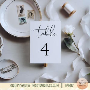 May include: White table number card with black calligraphy that reads "Table 4". The card is on a white surface with white fabric, a white flower, and a plate with vintage stamps.