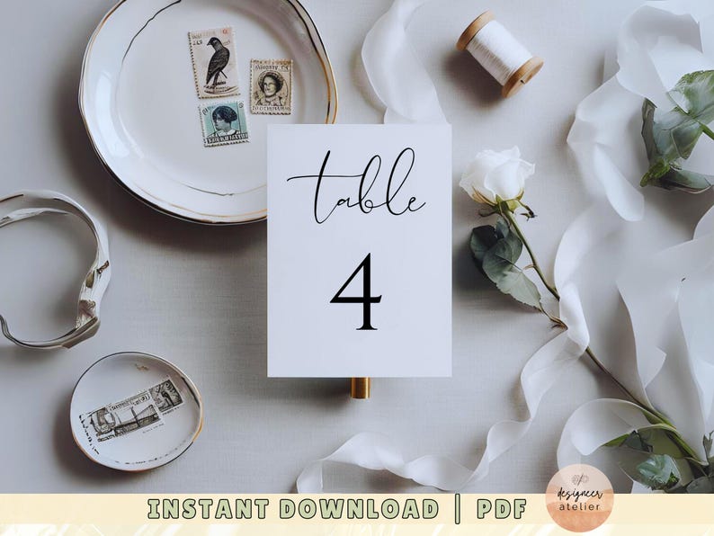 May include: White table number card with black calligraphy that reads "Table 4". The card is on a white surface with white fabric, a white flower, and a plate with vintage stamps.