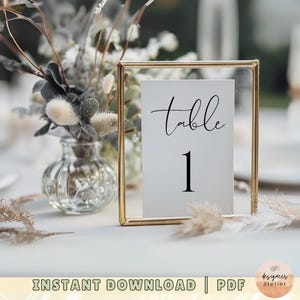 May include: A gold-framed table number card with the text "table 1" in black calligraphy. The card is sitting on a white table with dried flowers in a glass vase.
