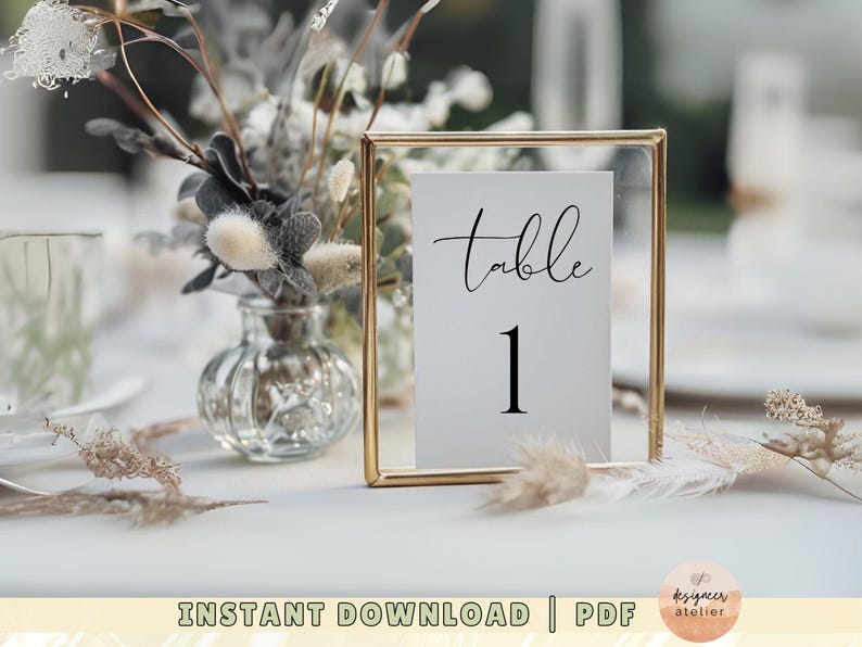 May include: A gold-framed table number card with the text "table 1" in black calligraphy. The card is sitting on a white table with dried flowers in a glass vase.