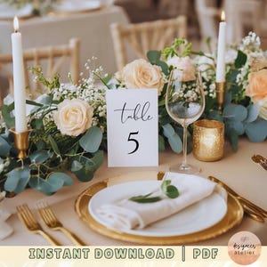 May include: A white card with black calligraphy that reads "table 5" sits on a table with a floral centerpiece. The centerpiece includes white and peach roses, greenery, and candles. A gold charger plate and white napkin are also on the table.