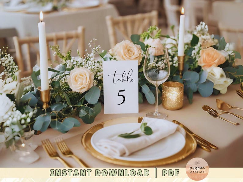 May include: A white card with black calligraphy that reads "table 5" sits on a table with a floral centerpiece. The centerpiece includes white and peach roses, greenery, and candles. A gold charger plate and white napkin are also on the table.