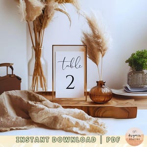 May include: A printable table number card with a gold frame and the text "Table 2" in black calligraphy. The card is sitting on a wooden table with dried pampas grass in vases.