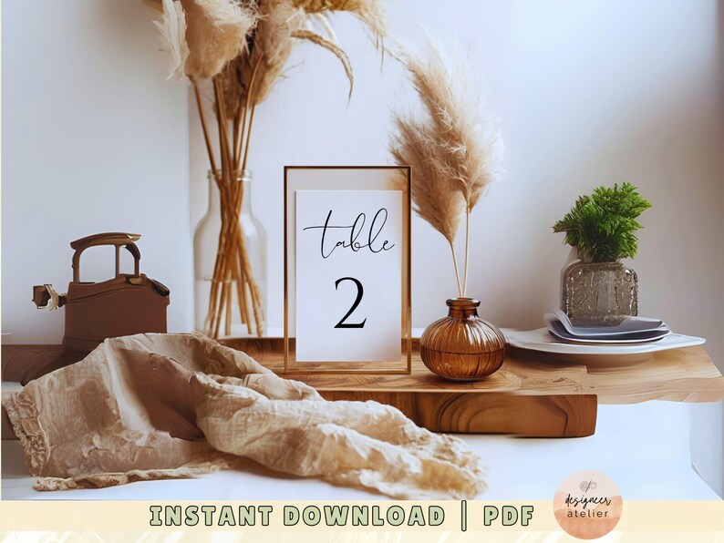 May include: A printable table number card with a gold frame and the text "Table 2" in black calligraphy. The card is sitting on a wooden table with dried pampas grass in vases.