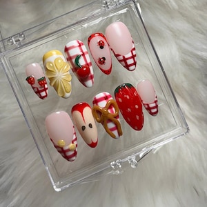 Puede incluir: Un conjunto de uñas postizas con temática de frutas. Los diseños incluyen fresas, limones, cerezas y manzanas. Algunas uñas presentan un patrón de cuadros rojos y blancos, mariquitas y un lazo. Las uñas se muestran en una caja de plástico transparente.