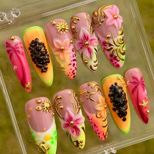 Puede incluir: Un conjunto de diez uñas artificiales con diversos diseños. Las uñas presentan una mezcla de colores rosa, naranja y verde, adornadas con detalles dorados y motivos florales y frutales en 3D. Las uñas están en una caja de plástico transparente.