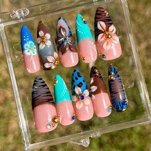 Puede incluir: Un conjunto de diez uñas artificiales en una caja de plástico transparente. Cada uña presenta un diseño diferente, incluyendo patrones florales, estampados de animales y arte abstracto, con una base rosa y varios colores de acento. Algunas uñas tienen adornos dorados o de joyas.