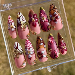 Puede incluir: Un conjunto de diez uñas artificiales en una caja de plástico transparente. Las uñas presentan varios diseños, incluyendo patrones florales, estampados de animales y detalles dorados. El color base es rosa claro, con detalles en blanco, burdeos y dorado.