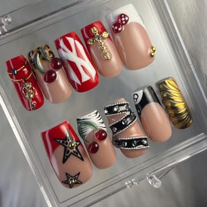May include: Set of ten false nails with various designs. The nails are red, white, black, and gold with cherries, stars, a cross, a baseball logo, and a zebra print.
