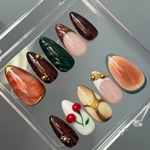 Puede incluir: Conjunto de diez uñas postizas con varios diseños. Las uñas son de forma almendrada y presentan una variedad de colores y patrones, incluyendo marrón, verde, blanco, dorado y rosa. Algunas uñas tienen detalles dorados y pequeñas cuentas doradas.