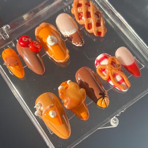 May include: A collection of custom-designed acrylic nails with various food-themed decorations. The nails feature designs such as a pie, a waffle, and a candy cane. The color palette includes shades of brown, orange, red, and white.