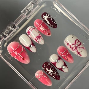 Könnte beinhalten: Ein Set mit zehn falschen Nägeln in einem durchsichtigen Plastiketui. Die Nägel sind lang und oval geformt. Sie sind mit einem Farbverlauf von Rosa und Burgunderrot lackiert, mit weißen Designs und Strasssteinen.