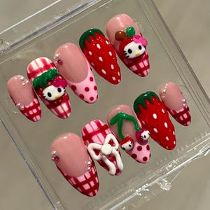 Puede incluir: Un conjunto de uñas postizas con varios diseños. Algunas uñas tienen una base rosa con cuadros rojos y blancos, mientras que otras presentan un diseño de fresa con lunares blancos. Algunas uñas tienen un diseño de Hello Kitty.