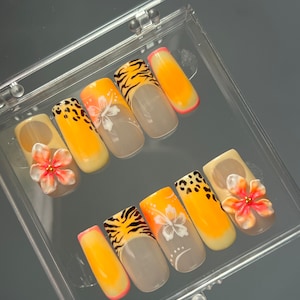 May include: A set of press-on nails in a clear plastic case. The nails feature various designs, including tiger stripes, leopard print, and floral accents. Colours include orange, yellow, pink, and white. Some nails have 3D flower embellishments.