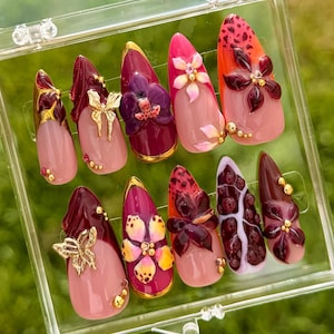 Puede incluir: Un conjunto de diez uñas artificiales en una caja de plástico transparente. Las uñas presentan una variedad de diseños, incluyendo detalles florales, adornos dorados y un estampado de leopardo. Los colores son rosa, rojo y dorado.