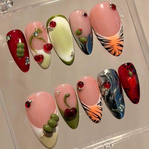 Puede incluir: Un conjunto de diez uñas artificiales en una caja de plástico transparente. Las uñas presentan varios diseños, incluyendo cerezas, aceitunas y rayas de tigre, con acentos de pedrería y un diseño de manicura francesa. Las uñas son de forma ovalada y están pintadas en tonos de rojo, verde, rosa y naranja.