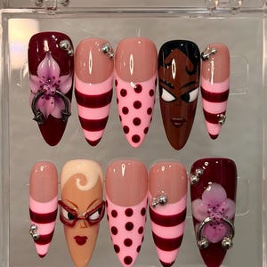 Puede incluir: Un juego de diez uñas artificiales en una caja de plástico transparente. Las uñas presentan varios diseños, incluyendo lunares rosas, rayas burdeos y una cara con gafas rojas. Algunas uñas tienen acentos florales y adornos plateados.