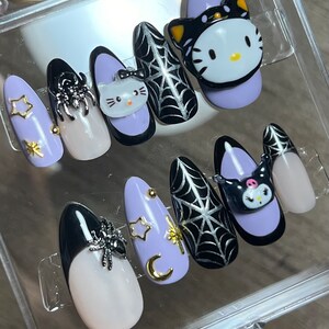 Puede incluir: Conjunto de diez uñas postizas con temática de Halloween. Las uñas están pintadas de un color morado claro con detalles en negro y plateado. Algunas uñas presentan una araña, una telaraña, una luna creciente y estrellas. Otras uñas presentan un gato negro con la cara de Hello Kitty.