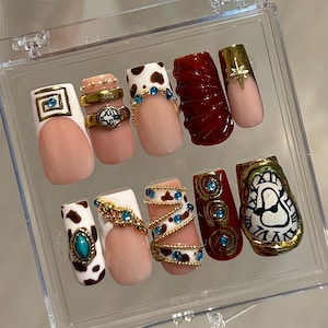 Puede incluir: Un conjunto de uñas postizas en una caja de plástico transparente. Las uñas presentan varios diseños, incluyendo estampado de vaca, detalles dorados y patrones intrincados. Los colores incluyen blanco, rosa, marrón, rojo y dorado. Algunas uñas tienen pedrería y anillos.