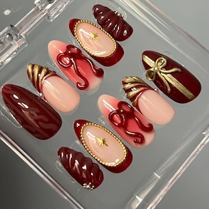 Puede incluir: Conjunto de diez uñas postizas con un diseño degradado en color burdeos y rosa. Cada uña presenta un acento dorado y un lazo rojo.