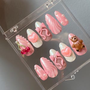 Puede incluir: Un conjunto de uñas postizas de color rosa y blanco con varios diseños, que incluyen un corazón, un lazo, una cereza y un osito de peluche.