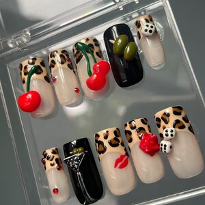 Op de afbeelding: Een set van tien kunstnagels met een luipaardprint. Elke nagel heeft een ander ontwerp, waaronder kersen, olijven, dobbelstenen, een martini-glas, een rode roos en een rode lip.