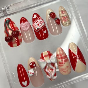 Puede incluir: Conjunto de diez uñas postizas de temática de San Valentín en rojo y blanco. Las uñas presentan una variedad de diseños que incluyen cerezas, lazos, corazones y un mensaje que dice "Te amo".