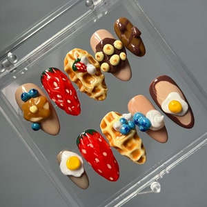 Puede incluir: Un conjunto de diez uñas postizas con temática de desayuno. Las uñas están decoradas con miniaturas de alimentos, incluyendo fresas, gofres, arándanos, chocolate y huevos.