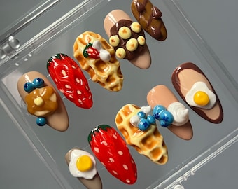 Peut inclure: Un ensemble de dix faux ongles sur le thème du petit-déjeuner. Les ongles sont décorés de miniatures d'aliments, notamment des fraises, des gaufres, des myrtilles, du chocolat et des œufs.