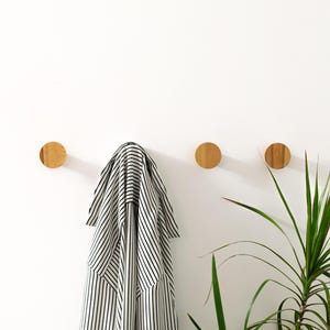 Op de afbeelding: Drie houten wandhaken met een gestreept shirt dat aan een van de haken hangt. Rechtsonder in de afbeelding staat een groene plant.