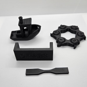 Puede incluir: Un conjunto de objetos negros impresos en 3D sobre una superficie blanca. Los objetos incluyen un pequeño barco, un engranaje circular, un marco rectangular con el texto "OF ABS" y una pieza en forma de pesa. Probablemente prototipos o modelos.