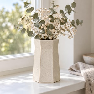 May include: A hexagonal ceramic vase with a speckled off-white finish containing a bouquet of dried eucalyptus branches and small white baby's breath flowers. The vase has a tapered neck and a wide base, standing upright on a smooth surface.