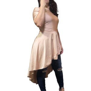 May include: A high-low blush pink top with a mock neck and short sleeves. The top has a flowing, asymmetrical hemline that is longer in the back and shorter in the front.