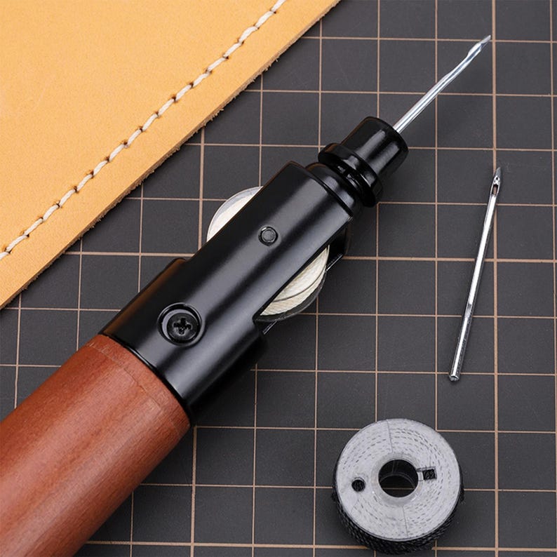 May include: A close-up of a leather sewing awl with a wooden handle and a black metal body. The awl has a small, round, white wheel on the side. The awl is lying on a black grid surface. There is a small, round, grey spool of thread next to the awl.