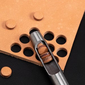 May include: A close-up of a leather punch tool with a stack of leather circles inside. The tool is made of metal and has a silver finish. The leather circles are brown and are used for making holes in leather.