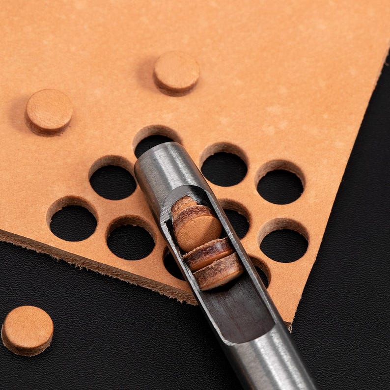 May include: A close-up of a leather punch tool with a stack of leather circles inside. The tool is made of metal and has a silver finish. The leather circles are brown and are used for making holes in leather.