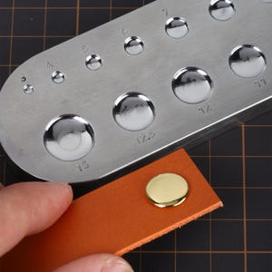 May include: A metal tool with 10 round holes of different sizes for punching leather. The tool is labeled with numbers 3, 4, 5, 6, 7, 11, 12, 12.5, 15. A gold-colored metal button is shown on a piece of brown leather.