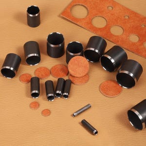 May include: A collection of cylindrical metal hollow punches in various diameters with sharpened edges, accompanied by several circular tan leather cutouts and a rectangular tan leather strip featuring multiple circular holes of different sizes.