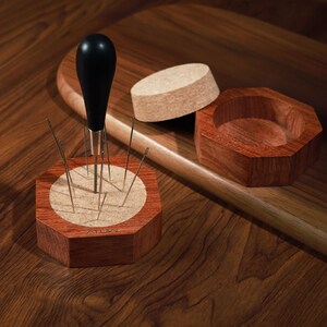 Puede incluir: Un port agujas de madera con agujas, un punz&oacute;n con mango negro, un corcho y un recipiente de madera. El port agujas es octogonal con un centro de color claro. El punz&oacute;n tiene un v&aacute;stago plateado.