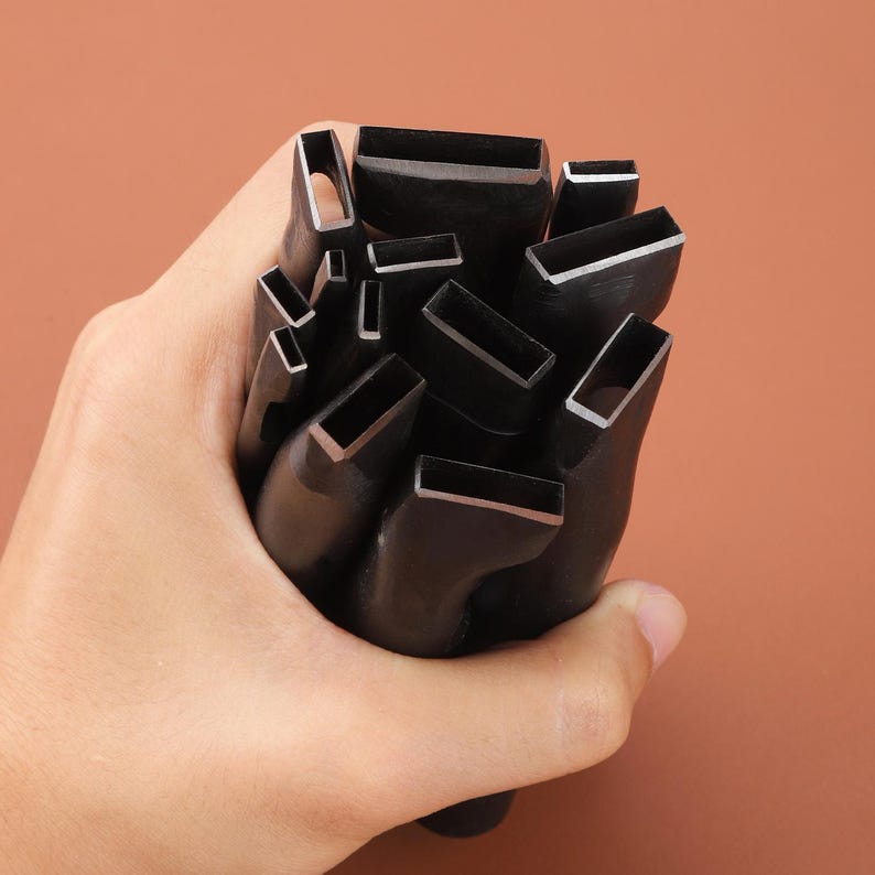 May include: A collection of black leathercraft tools with rectangular cutting heads. The tools are held in a hand against a brown background. The cutting edges have a silver finish.