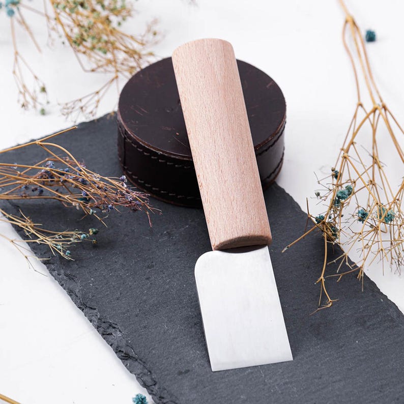 May include: A small, silver, curved blade with a wooden handle. The blade is on a black slate surface with a brown leather circle behind it.